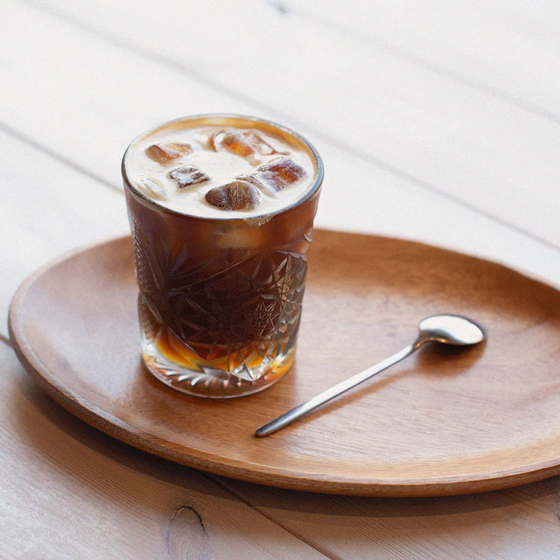 Szklanka mrożonej kawy z mlekiem i lodem na drewnianej tacy z łyżeczką – minimalistyczne ujęcie napoju.