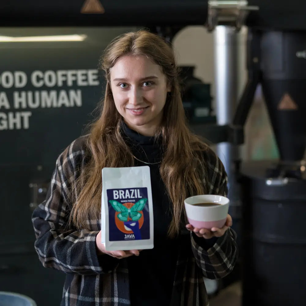 Kobieta w ciemnym płaszczu trzyma filiżankę z czarną kawą oraz opakowanie kawy Brazil Naimeg Paraiso od JAVA Coffee Roasters na tle palarni kawy. Rzemieślnicza kawa specialty z Brazylii, idealna do metod alternatywnych. W tle widoczny napis "Good Coffee is a Human Right".