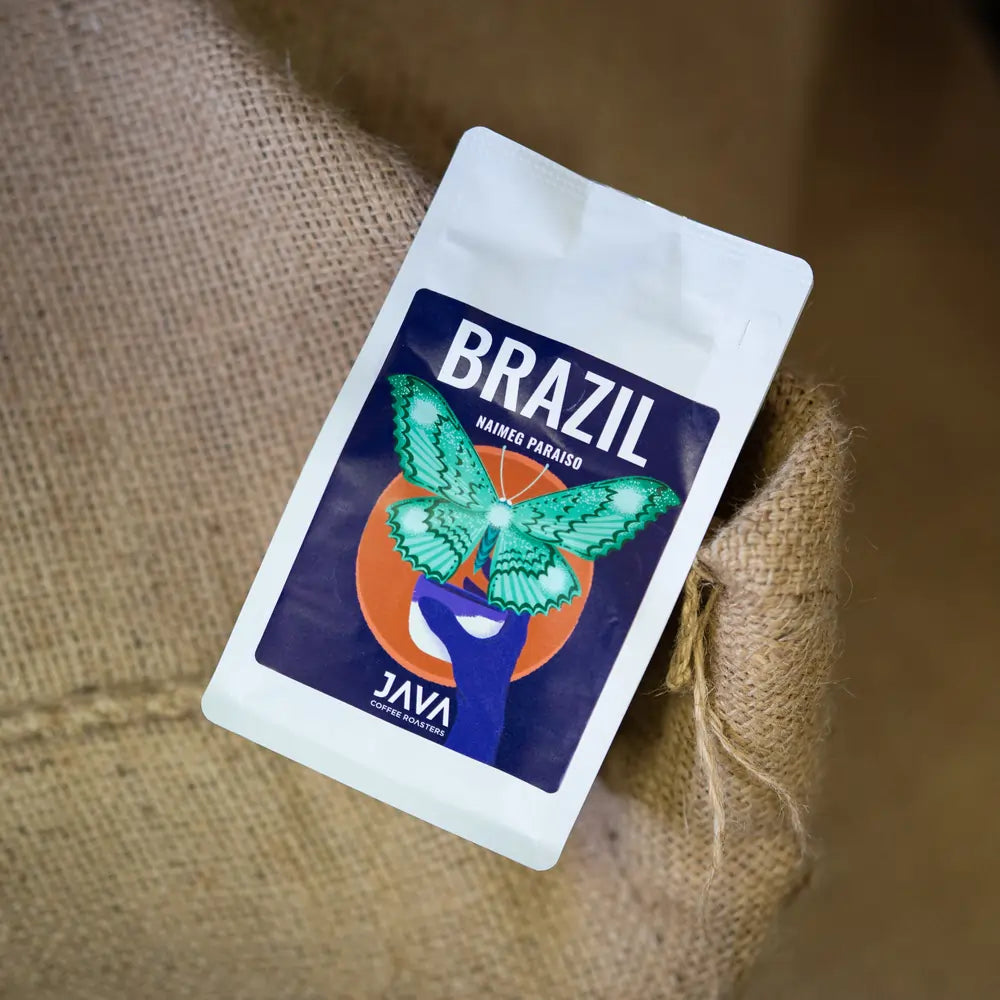 Opakowanie kawy Brazil Naimeg Paraiso od JAVA Coffee Roasters oparte na worku z ziarnami kawy. Świeżo palona kawa specialty z Brazylii, idealna do metod przelewowych.