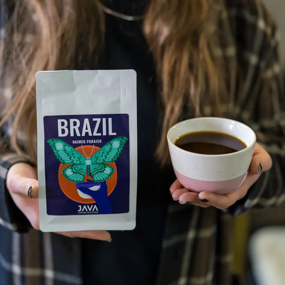 Osoba w ciemnym płaszczu trzyma filiżankę z czarną kawą oraz opakowanie kawy Brazil Naimeg Paraiso od JAVA Coffee Roasters. Świeżo palona kawa specialty z Brazylii, idealna do metod alternatywnych.