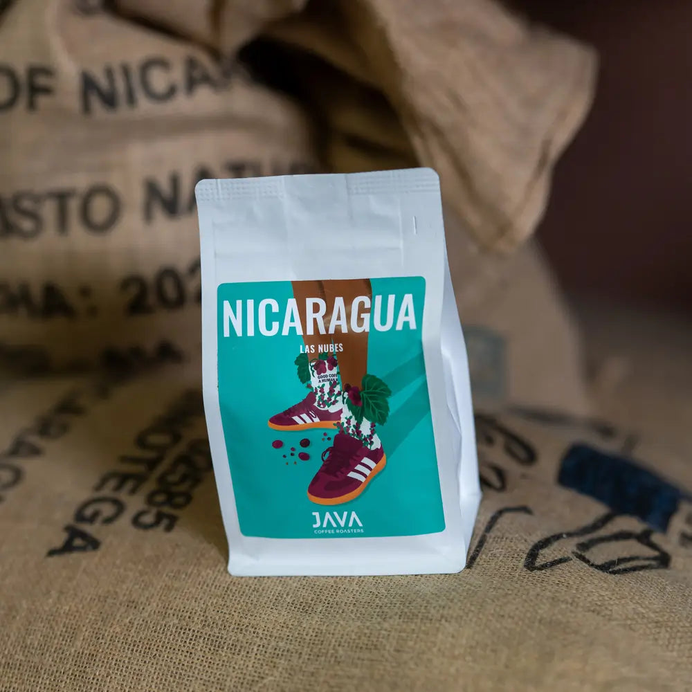 Opakowanie kawy Nicaragua Las Nubes od JAVA Coffee Roasters, ustawione na tle worków z zielonym ziarnem kawy. Etykieta przedstawia ilustrację butów wśród liści i owoców kawowca na turkusowym tle.