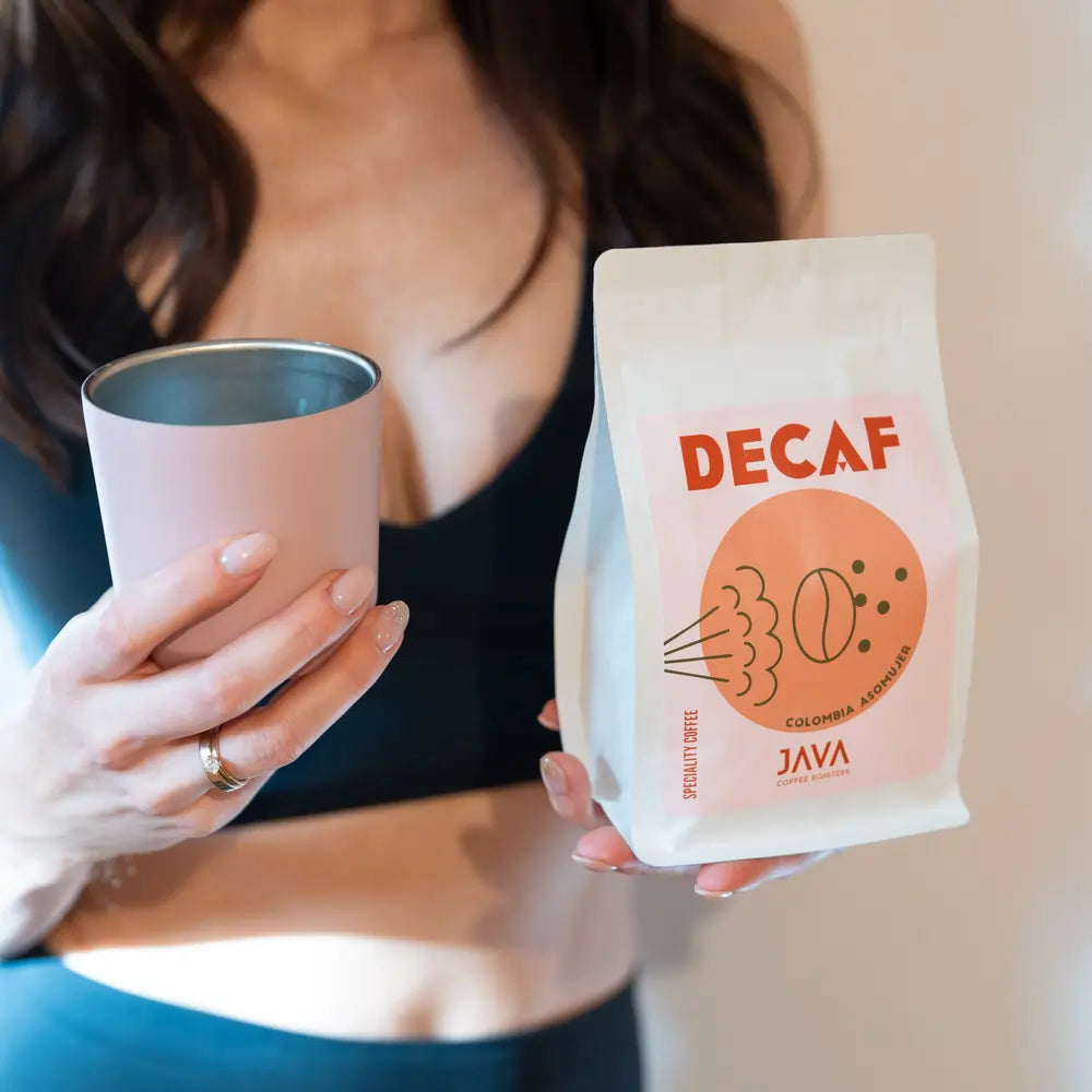 Kawa bezkofeinowa Java Coffee Roasters Decaf Colombia Asomuier 250g trzymana w dłoni obok kubka z czarną kawą