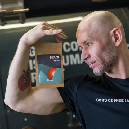 Mężczyzna w koszulce "Good Coffee is a Human Right" prezentuje opakowanie kawy Brazil Cerrado od JAVA Coffee Roasters, opierając je na swoim ramieniu. Palarz kawy w tle palarni. Świeżo palona kawa specialty z Brazylii, idealna do espresso.