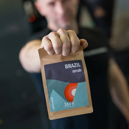 Mężczyzna w palarni kawy trzyma wyciągniętą ręką opakowanie Brazil Cerrado od JAVA Coffee Roasters. Kawa specialty z Brazylii, świeżo palona, idealna do espresso.