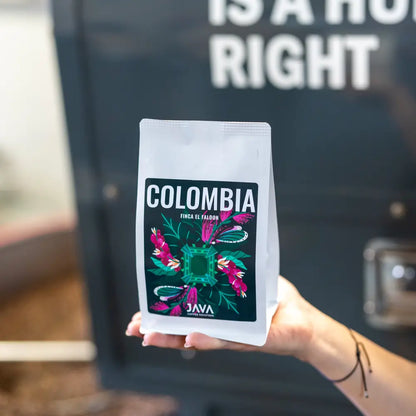 Opakowanie kawy specialty JAVA Coffee trzymane w dłoni na tle ciemnej powierzchni z białym napisem. Etykieta z napisem 'Colombia' i kolorową grafiką inspirowaną naturą. 