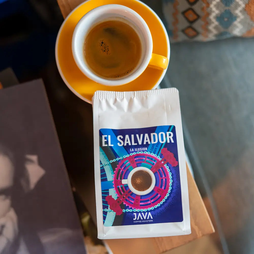 Filiżanka espresso w żółtym spodku obok paczki kawy ziarnistej El Salvador La Ilusion od Java Coffee Roasters, leżącej na drewnianym stoliku obok książki i poduszki z etnicznym wzorem