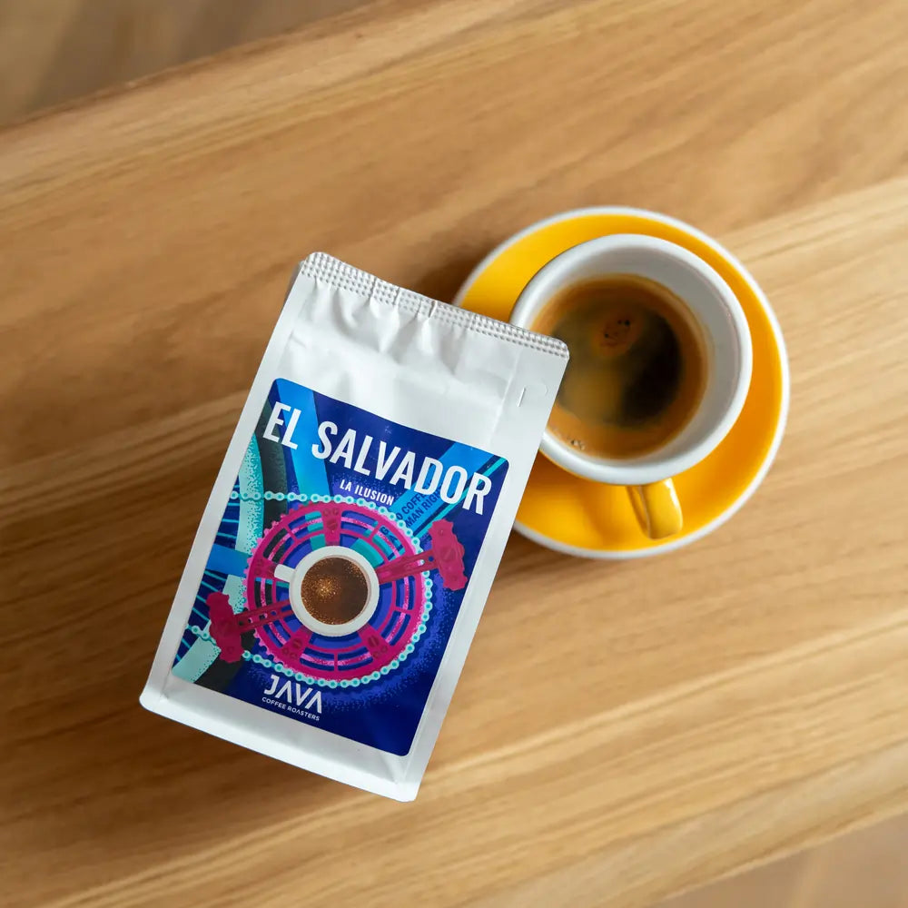 Filiżanka espresso w żółtym spodku obok paczki kawy ziarnistej El Salvador La Ilusion od Java Coffee Roasters, leżącej na drewnianym stoliku