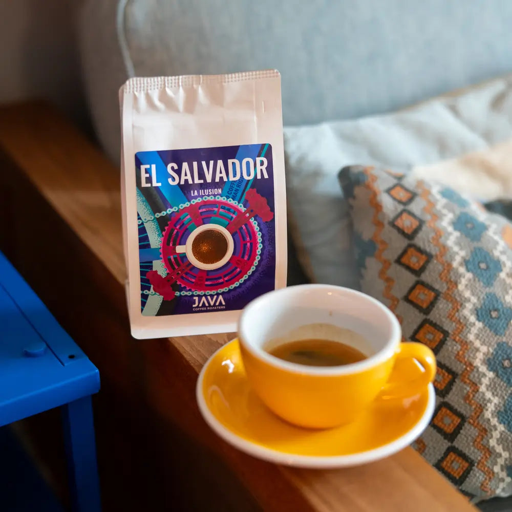 Filiżanka espresso w żółtym spodku obok paczki kawy ziarnistej El Salvador La Ilusion od Java Coffee Roasters, stojącej na drewnianym stoliku obok książki i poduszki z etnicznym wzorem