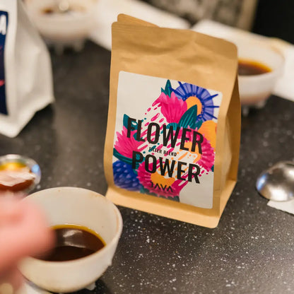 Paczka kawy Flower Power od JAVA Coffee Roasters na czarnym blacie podczas cuppingu, otoczona filiżankami z zaparzoną kawą i akcesoriami.