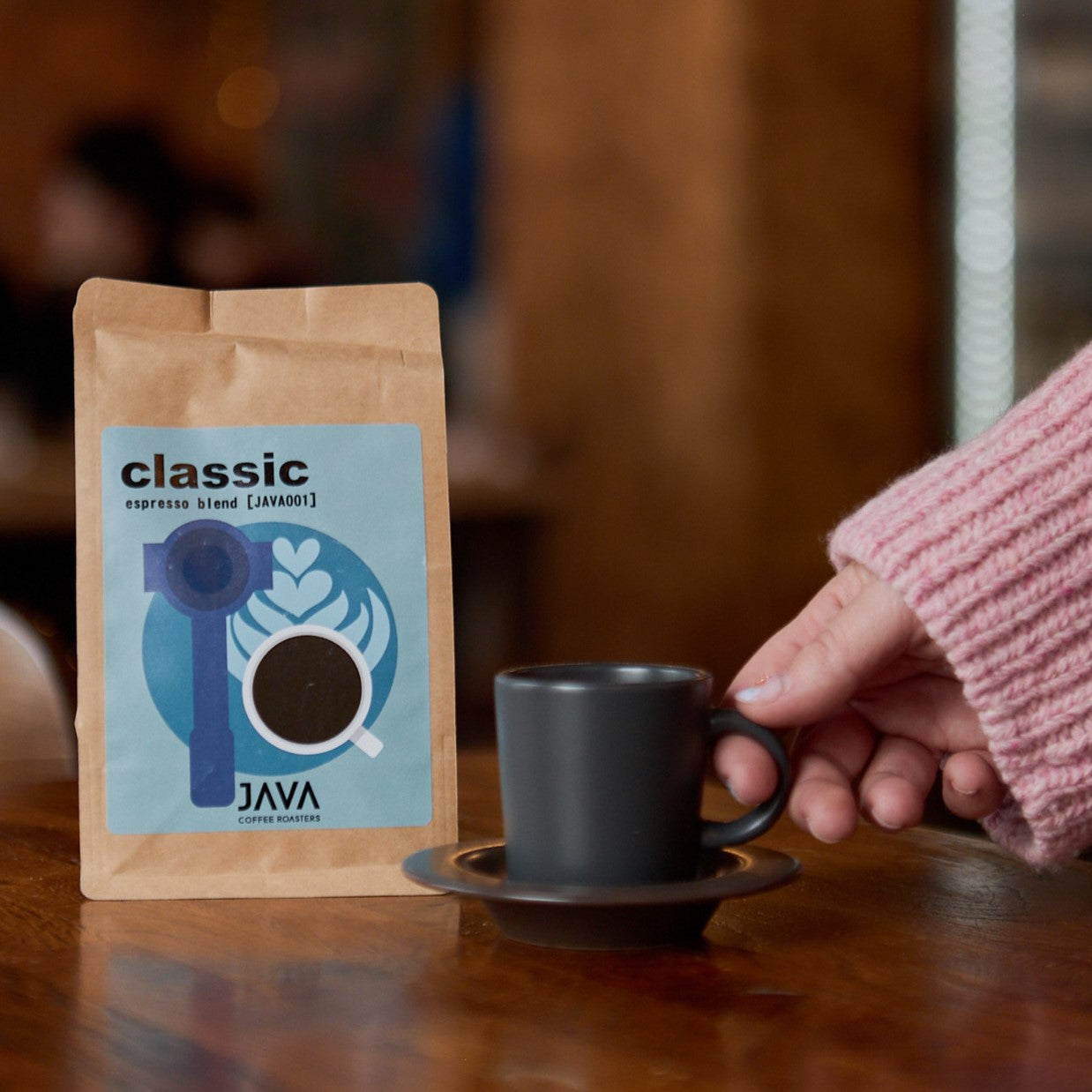 Opakowanie kawy JAVA Coffee Roasters "Classic" (espresso blend) z niebieską etykietą przedstawiającą portafilter i filiżankę z kawą oraz wzorem latte art. Osoba w różowym swetrze trzyma czarną filiżankę unoszoną nad spodkiem na drewnianym stole. W tle przytulne wnętrze kawiarni z ciepłym oświetleniem.