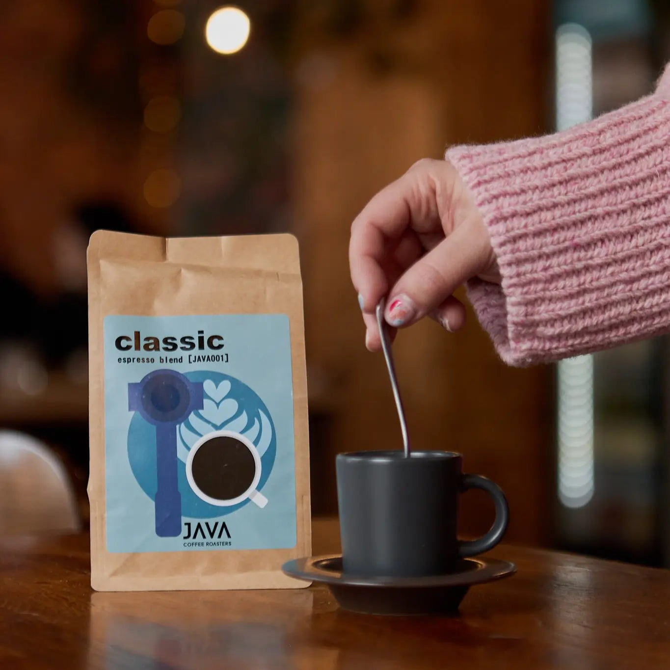 Opakowanie kawy JAVA Coffee Roasters "Classic" (espresso blend) z niebieską etykietą, przedstawiającą portafilter i filiżankę z kawą oraz wzorem latte art. Obok czarna filiżanka na spodku, a osoba w różowym swetrze miesza kawę łyżeczką. W tle przytulne wnętrze kawiarni z ciepłym oświetleniem.