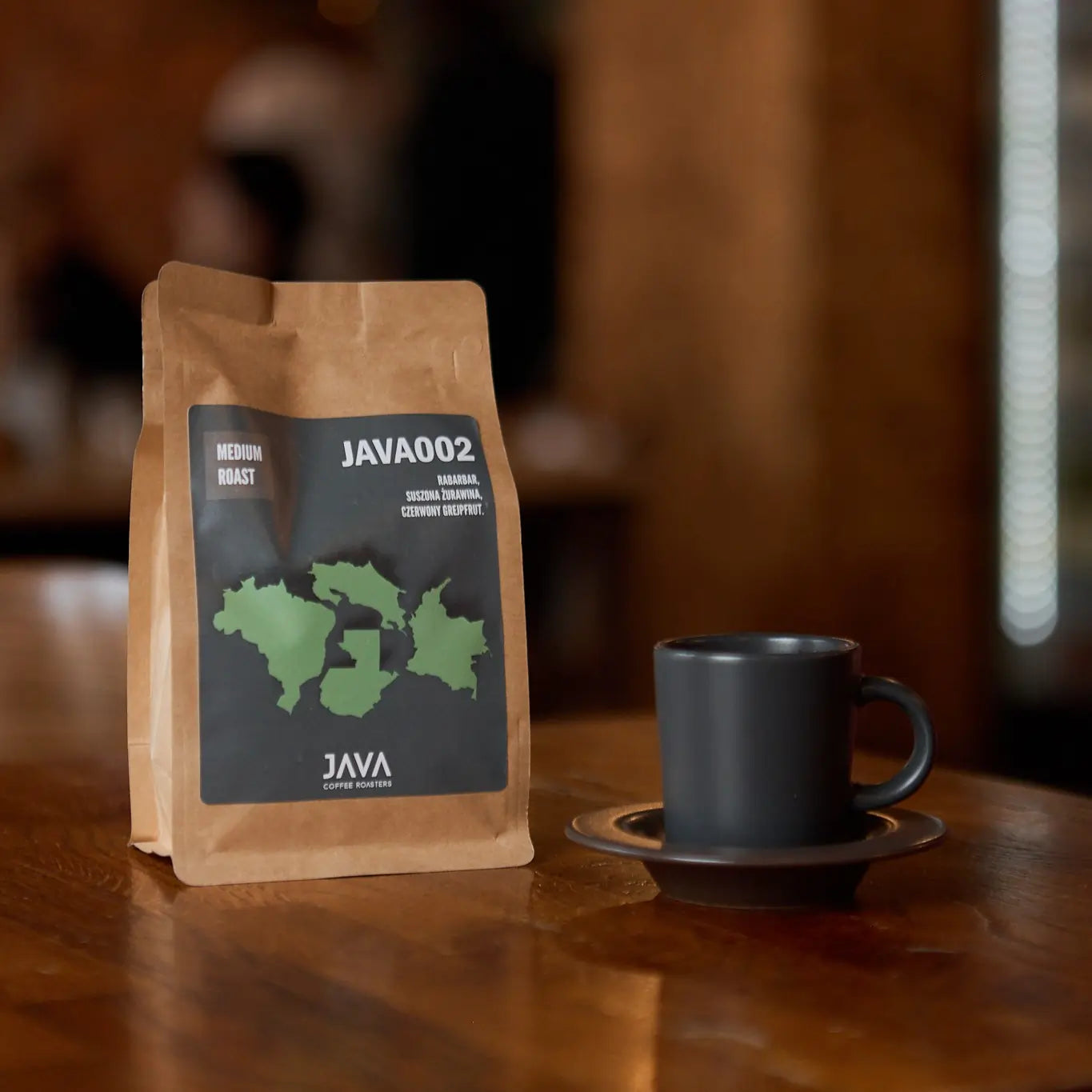 Opakowanie kawy JAVA Coffee Roasters "JAVA002" o średnim stopniu palenia, z czarno-zieloną etykietą przedstawiającą zarys mapy regionów. Na opakowaniu widoczne są nuty smakowe: rabarbar, suszona żurawina, czerwony grejpfrut. Obok na drewnianym stole stoi czarna filiżanka na spodku, a w tle widać rozmyte, ciepłe światło wnętrza kawiarni.