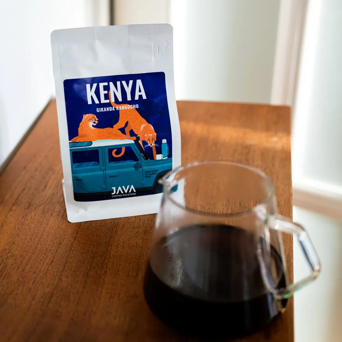 Kawa ziarnista Kenya Gikanda Kangoch od JAVA Coffee Roasters – opakowanie kawy na drewnianym stole obok zaparzonej czarnej kawy w szklanym dzbanku.