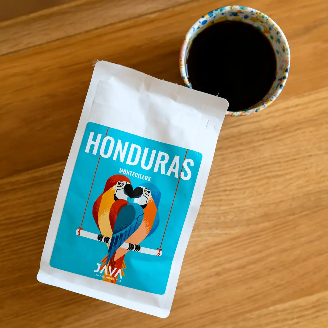 Opakowanie kawy Java Honduras Montecillos 250g obok filiżanki z zaparzoną kawą, ujęcie z góry na drewnianym stole