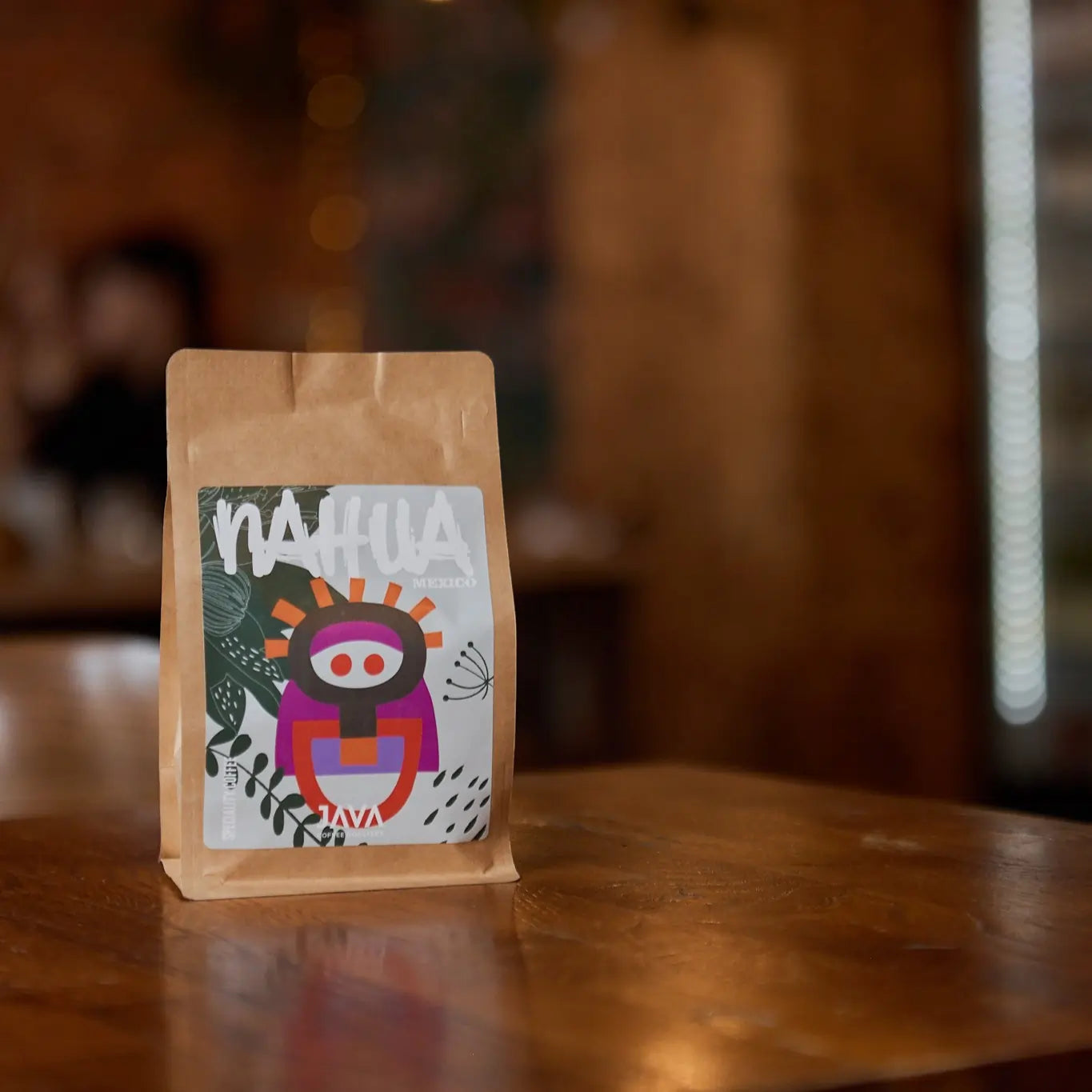 Opakowanie kawy JAVA Coffee Roasters "Nahua" z Meksyku, z kolorową grafiką inspirowaną sztuką ludową na szaro-zielonym tle z roślinnymi motywami. Opakowanie stoi na drewnianym stole w przytulnym wnętrzu z rozmytym, ciepłym światłem w tle.