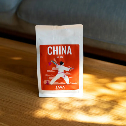 Opakowanie kawy JAVA Coffee Roasters China Gui Ben stojące na drewnianym stole w promieniach słońca, z rozproszonym światłem i miękkim tłem sofy.
