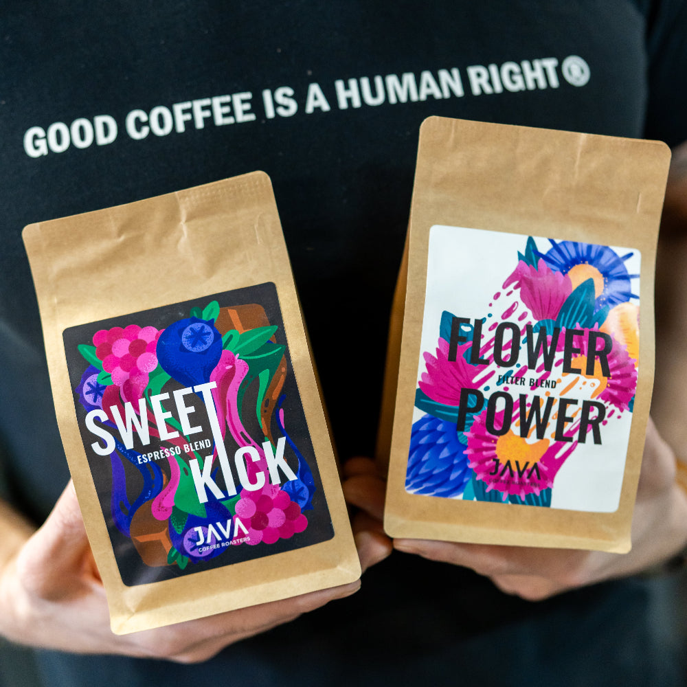  Kawa specialty Java Coffee Roasters – blendy Sweet Kick i Flower Power w brązowych torebkach z kolorowymi etykietami, trzymane w dłoniach osoby w koszulce z napisem „GOOD COFFEE IS A HUMAN RIGHT”.