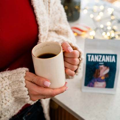 Kobieta trzyma kubek z kawą, a w tle stoi opakowanie JAVA Coffee Tanzania Itumpi w świątecznym wnętrzu.