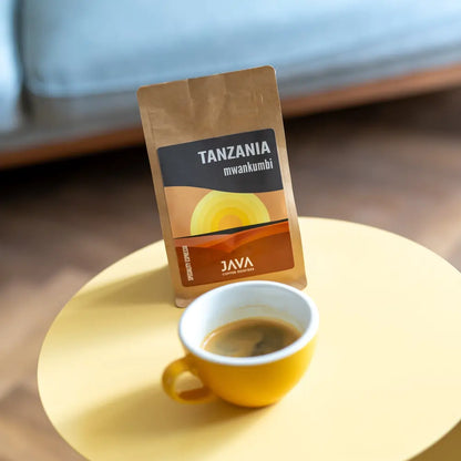 Torebka kawy Java Coffee Roasters "Tanzania Mwankumbi" na żółtym stoliku, obok filiżanki espresso w żółtej filiżance. W tle rozmazana kanapa i drewniana podłoga.