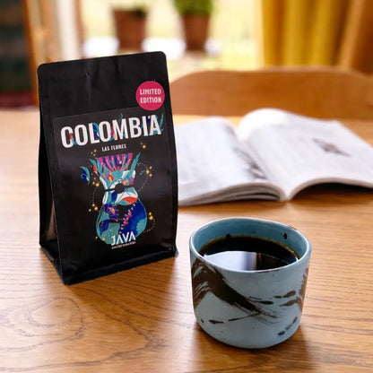 Kawa ziarnista Colombia Las Flores JAVA Coffee oraz filiżanka czarnej kawy z książką w tle na drewnianym stole