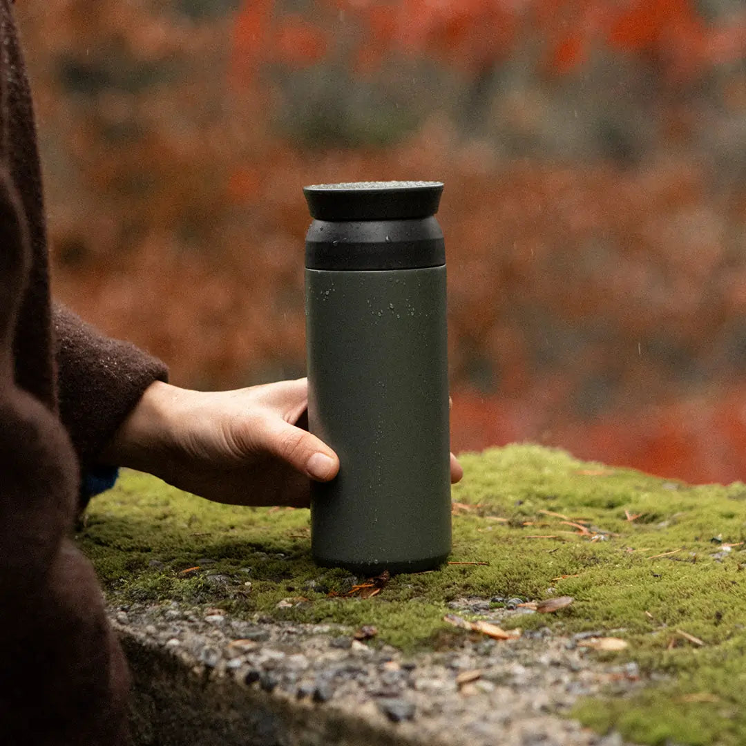 Kubek termiczny Kinto Travel Tumbler w kolorze Ash Green trzymany w dłoni na tle jesiennego krajobrazu – zdjęcie lifestyle.