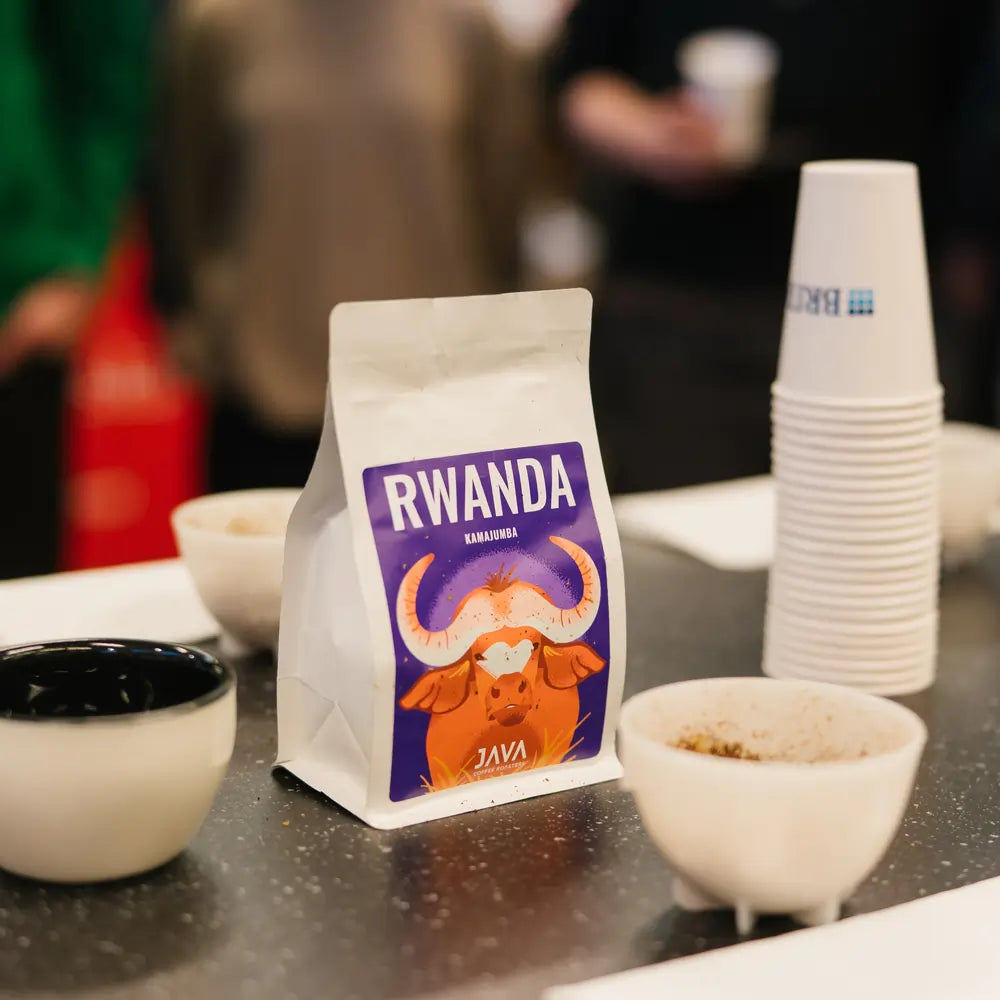 Paczka kawy Rwanda Kamajumba od JAVA Coffee Roasters ustawiona na blacie podczas cuppingu, obok filiżanek z kawą i stosu papierowych kubków, w tle uczestnicy degustacji