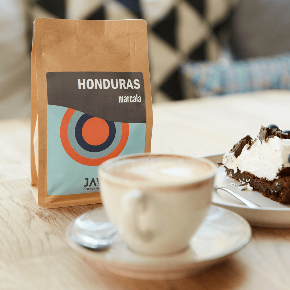 Brązowe opakowanie kawy JAVA Coffee Honduras Marcala na stole obok filiżanki cappuccino i kawałka ciasta czekoladowego z bitą śmietaną. Przytulne wnętrze kawiarni w tle