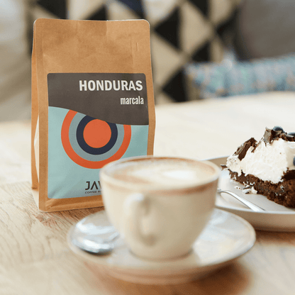 Brązowe opakowanie kawy JAVA Coffee Honduras Marcala na stole obok filiżanki cappuccino i kawałka ciasta czekoladowego z bitą śmietaną. Przytulne wnętrze kawiarni w tle