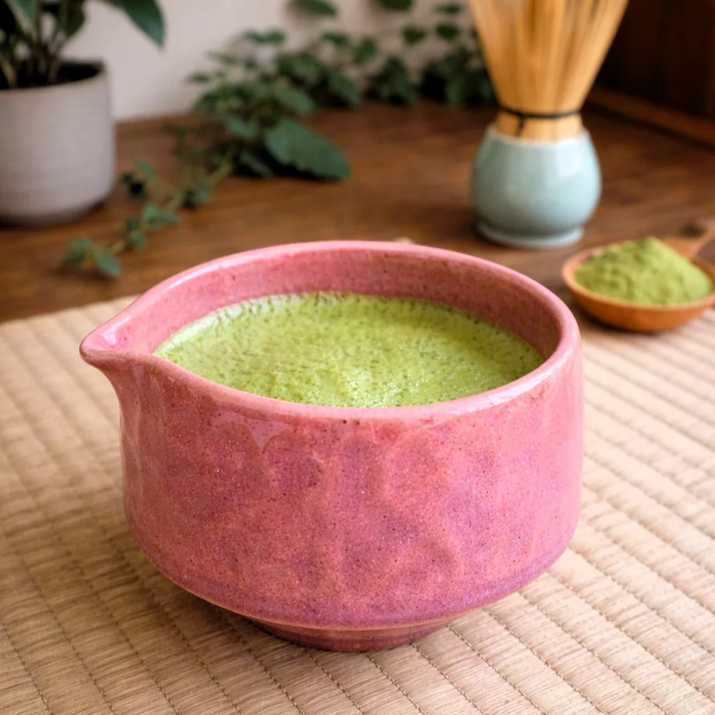 Różowa ceramiczna czarka do matchy z dziobkiem Matchawan Botan wypełniona zieloną matchą, ustawiona na macie w naturalnym, herbacianym otoczeniu.