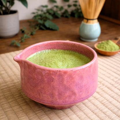 Różowa ceramiczna czarka do matchy z dziobkiem Matchawan Botan wypełniona zieloną matchą, ustawiona na macie w naturalnym, herbacianym otoczeniu.