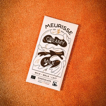 Tabliczka mlecznej czekolady Meurisse Creamy Milk 39% kakao z certyfikatami Fairtrade i Bio, sfotografowana na pomarańczowym tle.