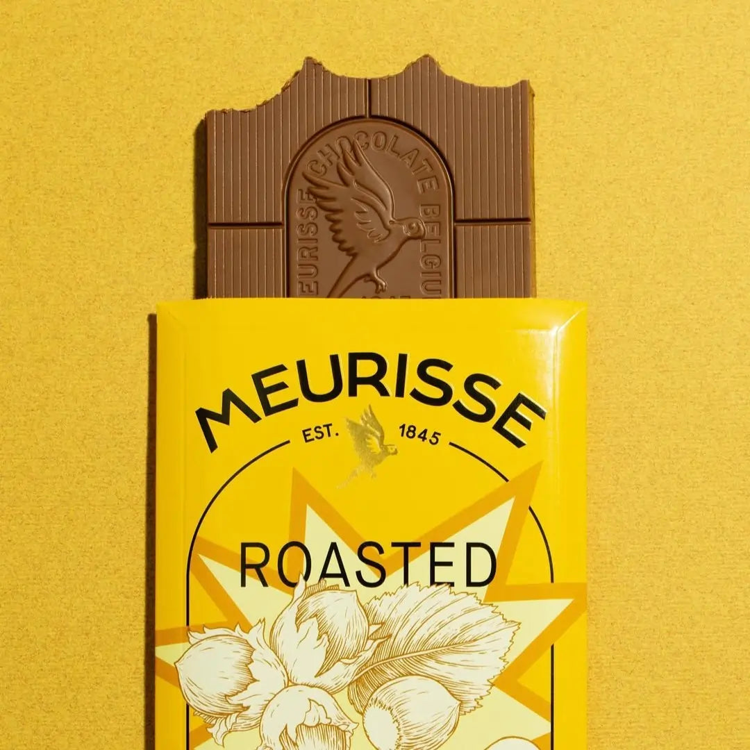 Tabliczka mlecznej czekolady Meurisse Roasted Hazelnuts 39% kakao częściowo wysunięta z żółtego opakowania z ilustracją orzechów laskowych, na tle w odcieniu żółtym.