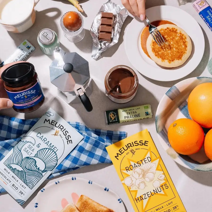 Śniadaniowa kompozycja z tabliczką mlecznej czekolady Meurisse Roasted Hazelnuts 39% kakao, Caramel & Sea Salt, kawą, naleśnikami i owocami na jasnym tle.