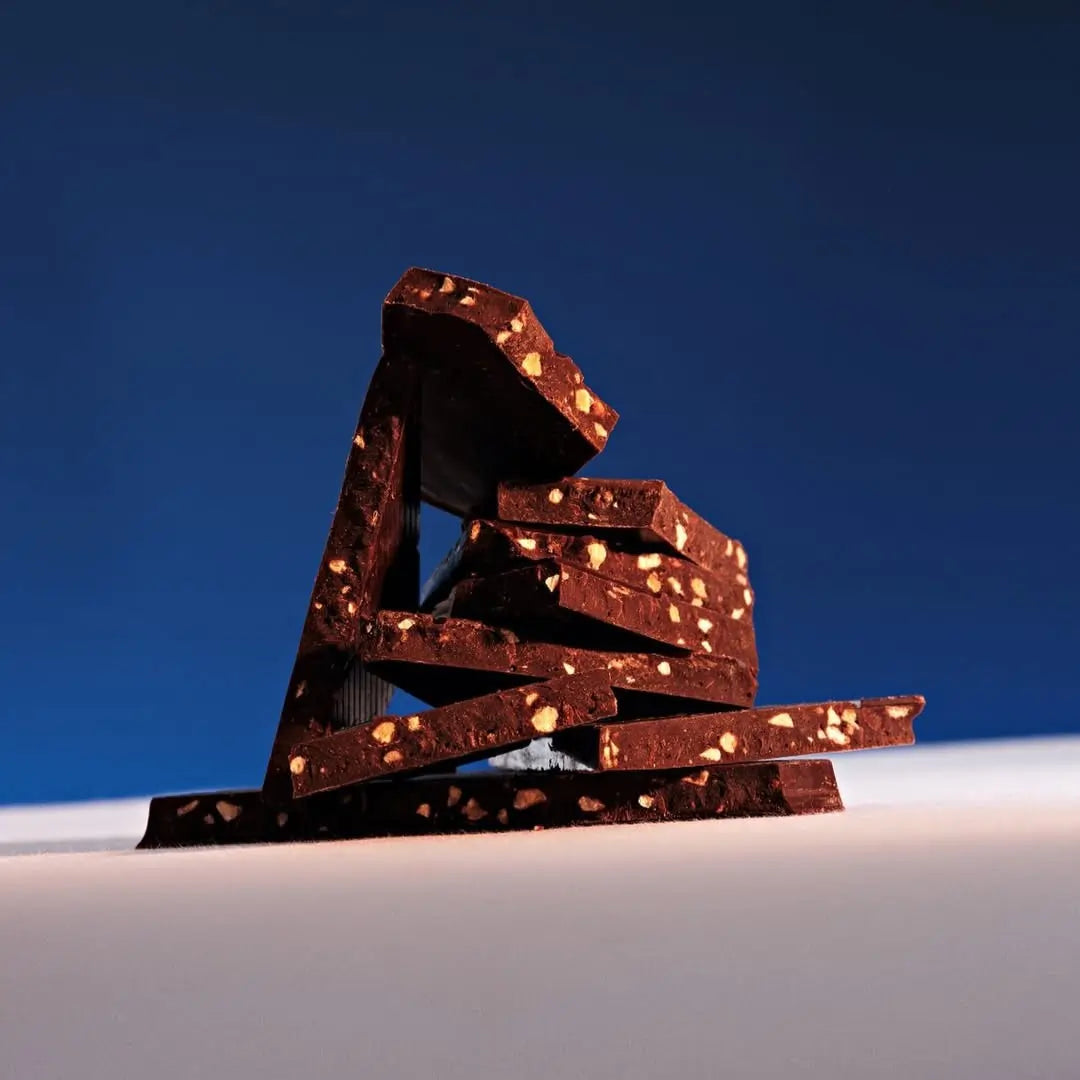Połamane kawałki mlecznej czekolady Meurisse z prażonymi orzechami laskowymi, ułożone w artystyczną kompozycję na tle granatowego nieba.