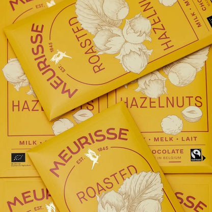 Kilka tabliczek mlecznej czekolady Meurisse Roasted Hazelnuts 39% kakao ułożonych płasko, w żółtych opakowaniach z ilustracją orzechów laskowych i certyfikatami BIO oraz Fairtrade.