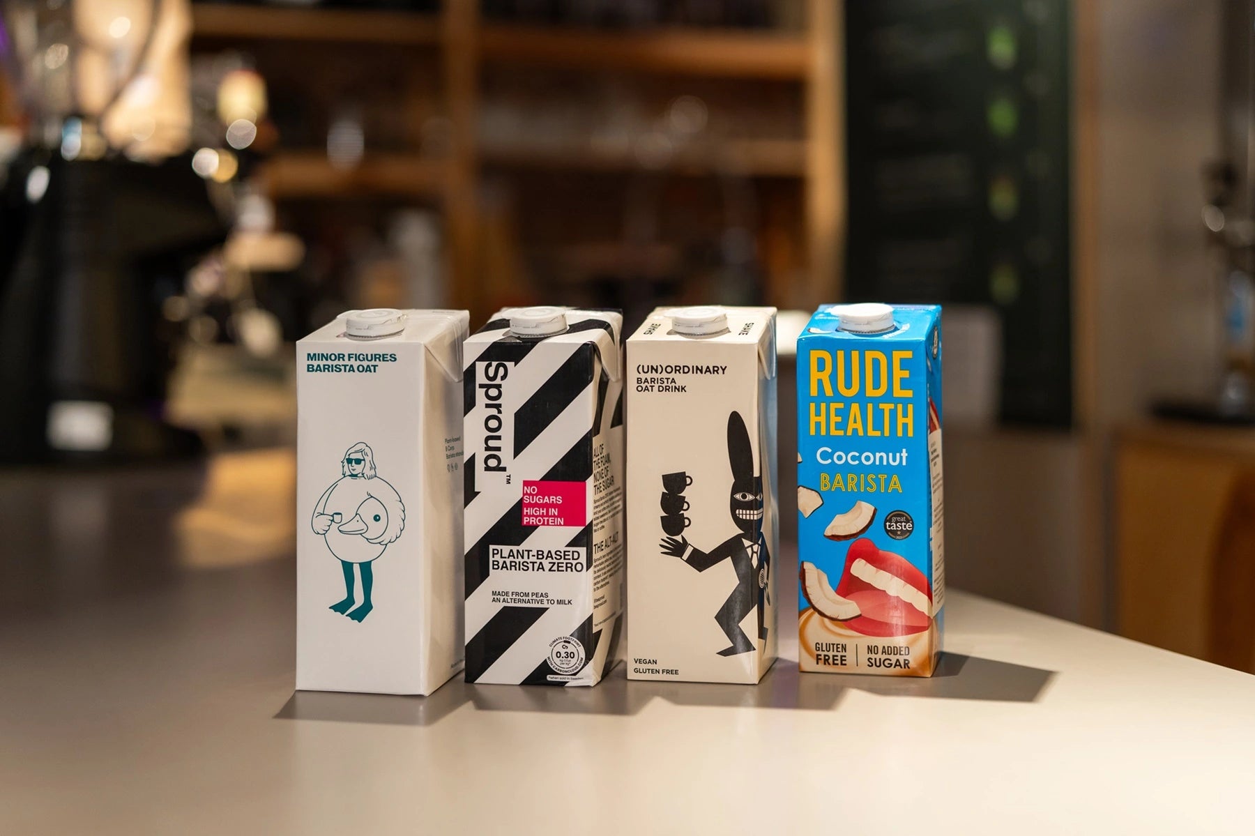 Zestaw napojów roślinnych dla baristów: Minor Figures Barista Oat, Sproud Plant-Based Barista Zero, (Un)Ordinary Barista Oat Drink oraz Rude Health Coconut Barista, ustawione na blacie w kawiarni.