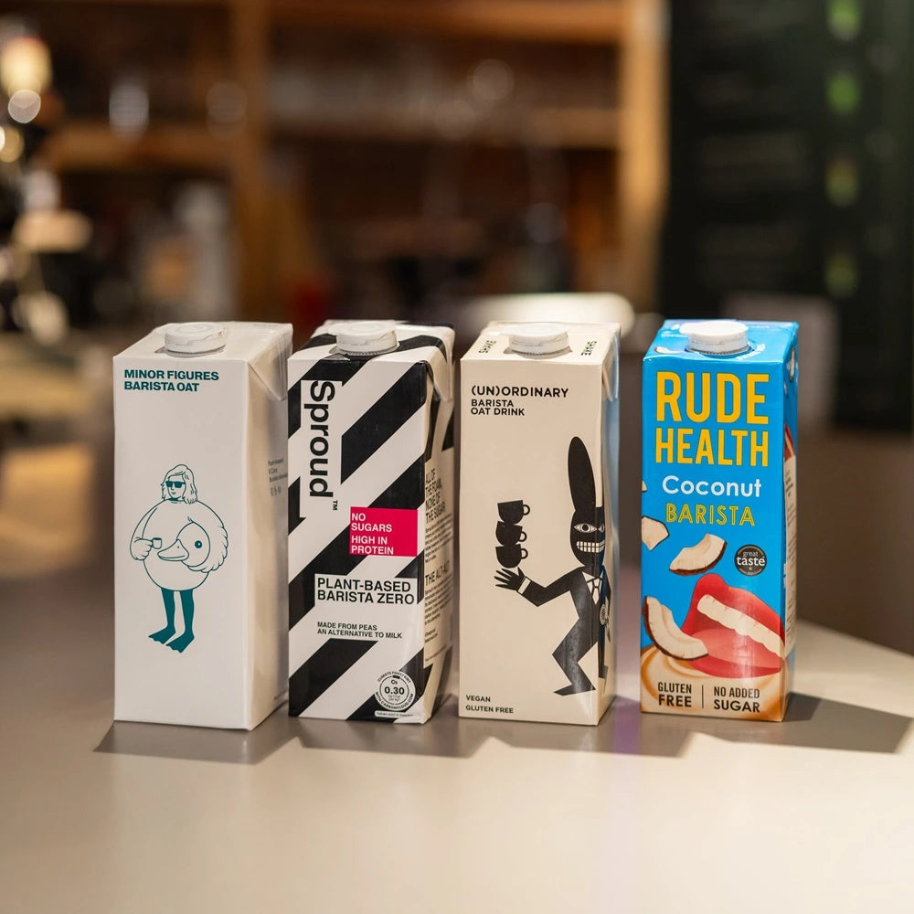 Zestaw napojów roślinnych dla baristów: Minor Figures Barista Oat, Sproud Plant-Based Barista Zero, (Un)Ordinary Barista Oat Drink oraz Rude Health Coconut Barista, ustawione na blacie w kawiarni