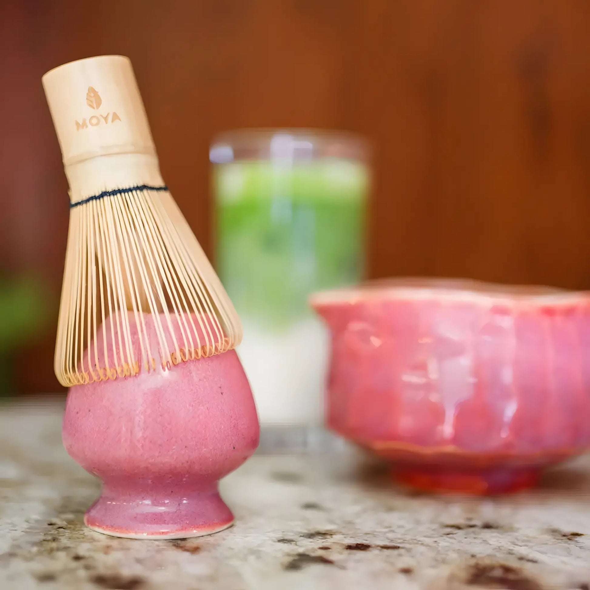 Różowa ceramiczna podstawka do trzepaczki do matchy (chanoy) z bambusową trzepaczką chasen, w tle czarka do matchy i szklanka latte matcha.