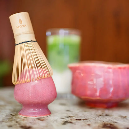 Różowa ceramiczna podstawka do trzepaczki do matchy (chanoy) z bambusową trzepaczką chasen, w tle czarka do matchy i szklanka latte matcha.