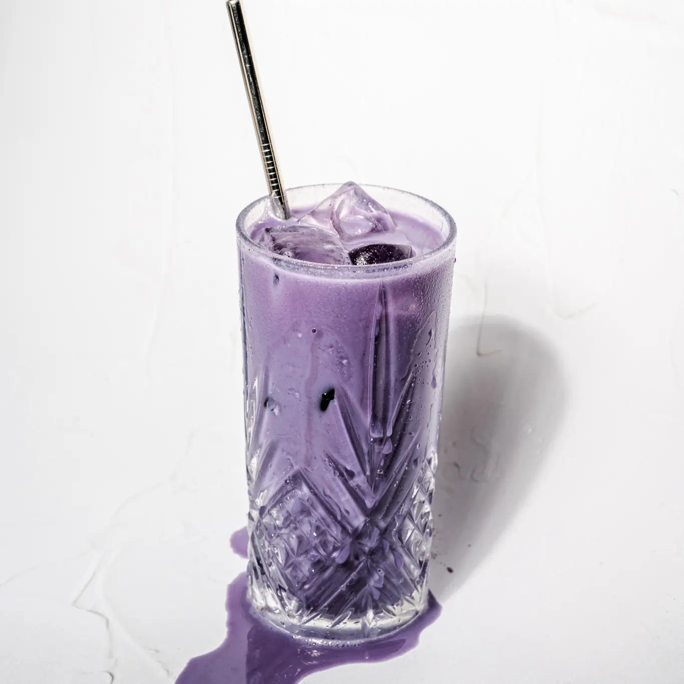 Mrożone fioletowe latte z ube i kostkami lodu podane w dekoracyjnej szklance ze słomką na jasnym tle
