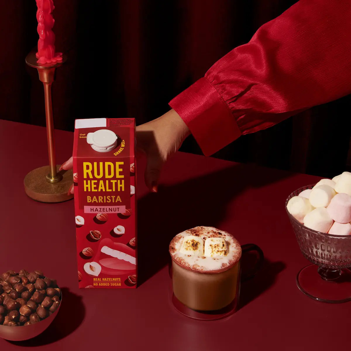 Napój roślinny Rude Health Barista Hazelnut 1L na bordowym tle, obok filiżanki gorącej czekolady z piankami, miseczki orzechów laskowych i zapalonej świecy – elegancka, ciepła kompozycja w odcieniach czerwieni.