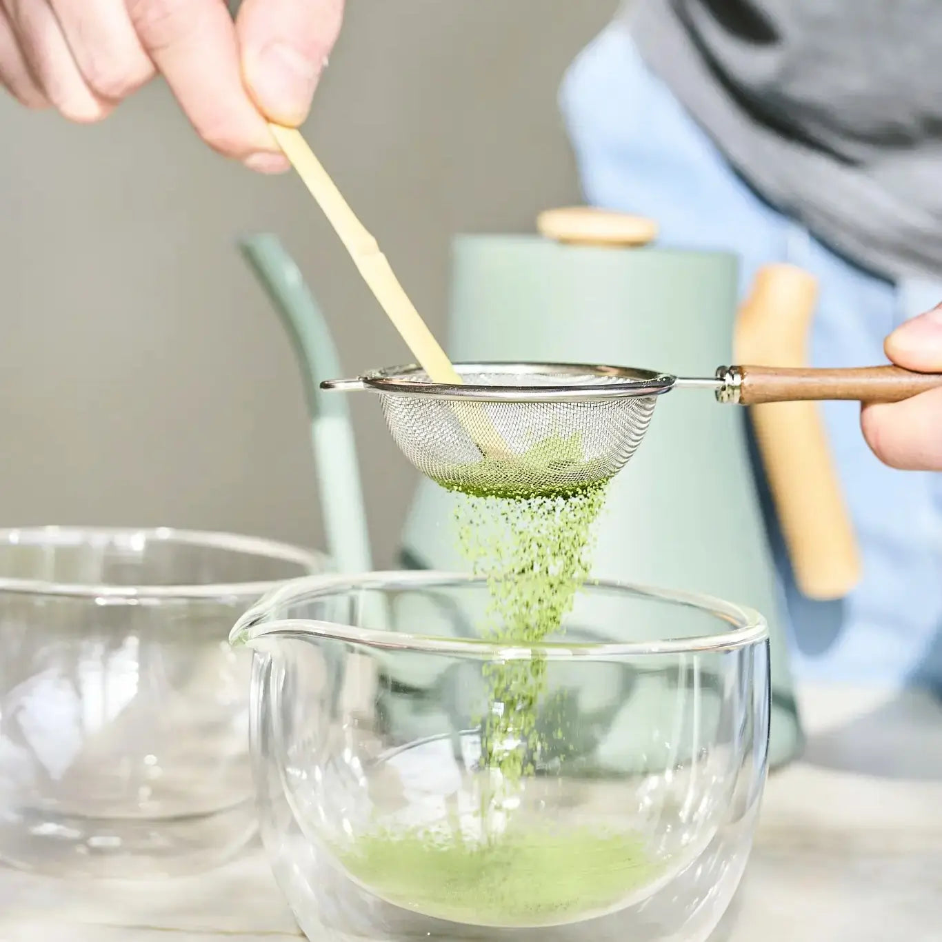 Stalowe sitko z drewnianą rączką podczas przesiewania zielonej matchy do szklanej czarki, przygotowanie herbaty matcha z użyciem bambusowej łyżeczki chashaku.