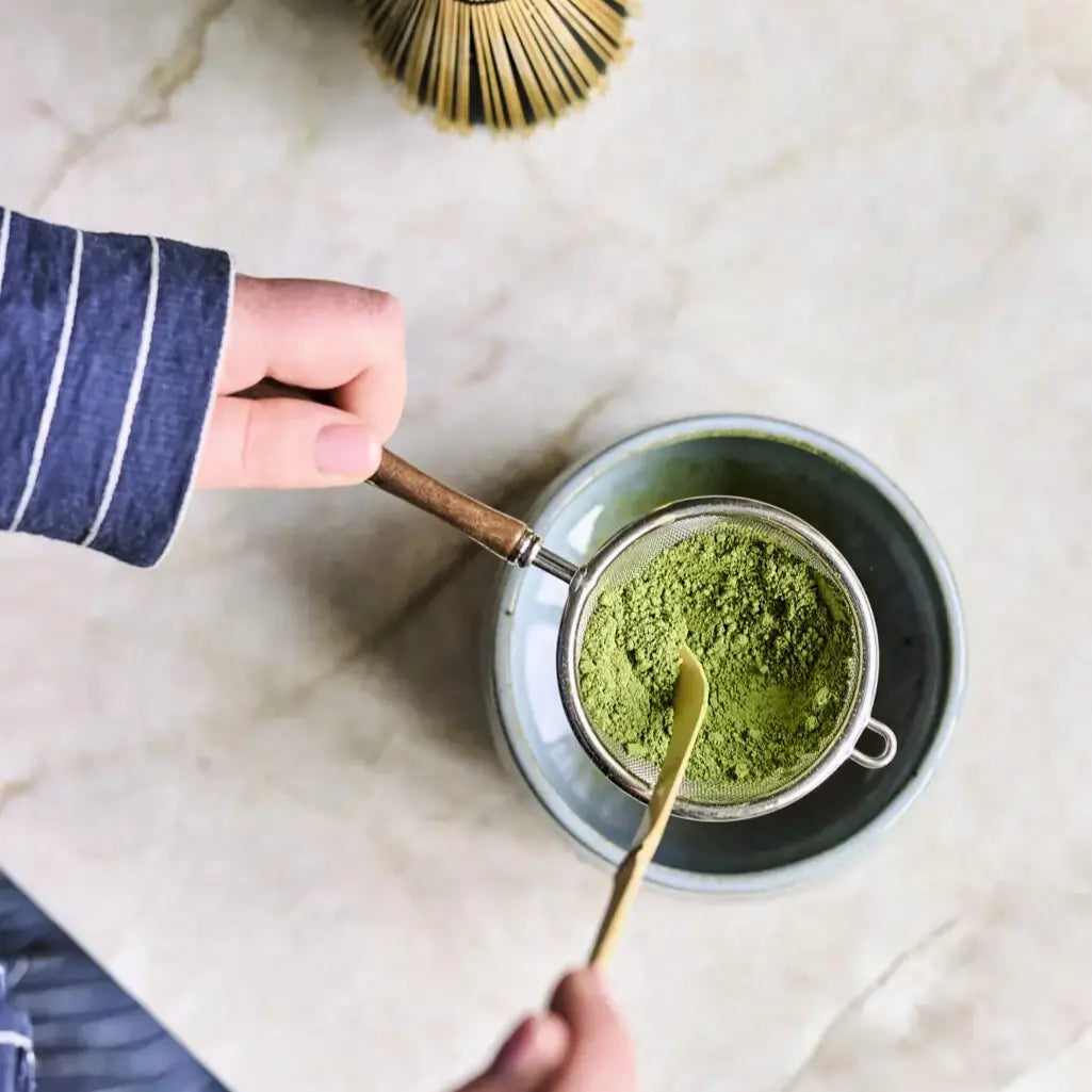 Stalowe sitko z drewnianą rączką do przesiewania matchy nad czarką, widok z góry, przygotowanie herbaty matcha z użyciem bambusowej łyżeczki chashaku.