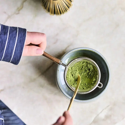 Stalowe sitko z drewnianą rączką do przesiewania matchy nad czarką, widok z góry, przygotowanie herbaty matcha z użyciem bambusowej łyżeczki chashaku.