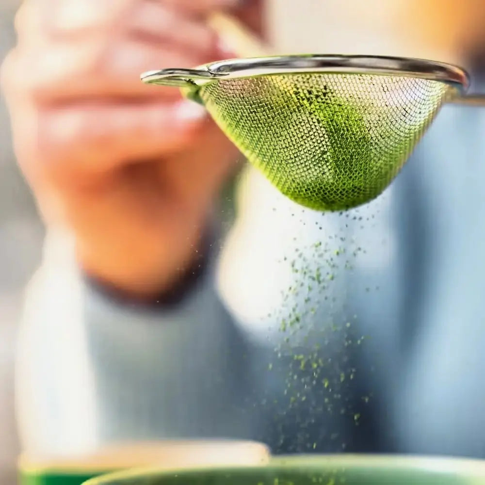 Stalowe sitko do matchy podczas przesiewania zielonego proszku matcha nad czarką, widoczny opadający pył i drobna siateczka w zbliżeniu.