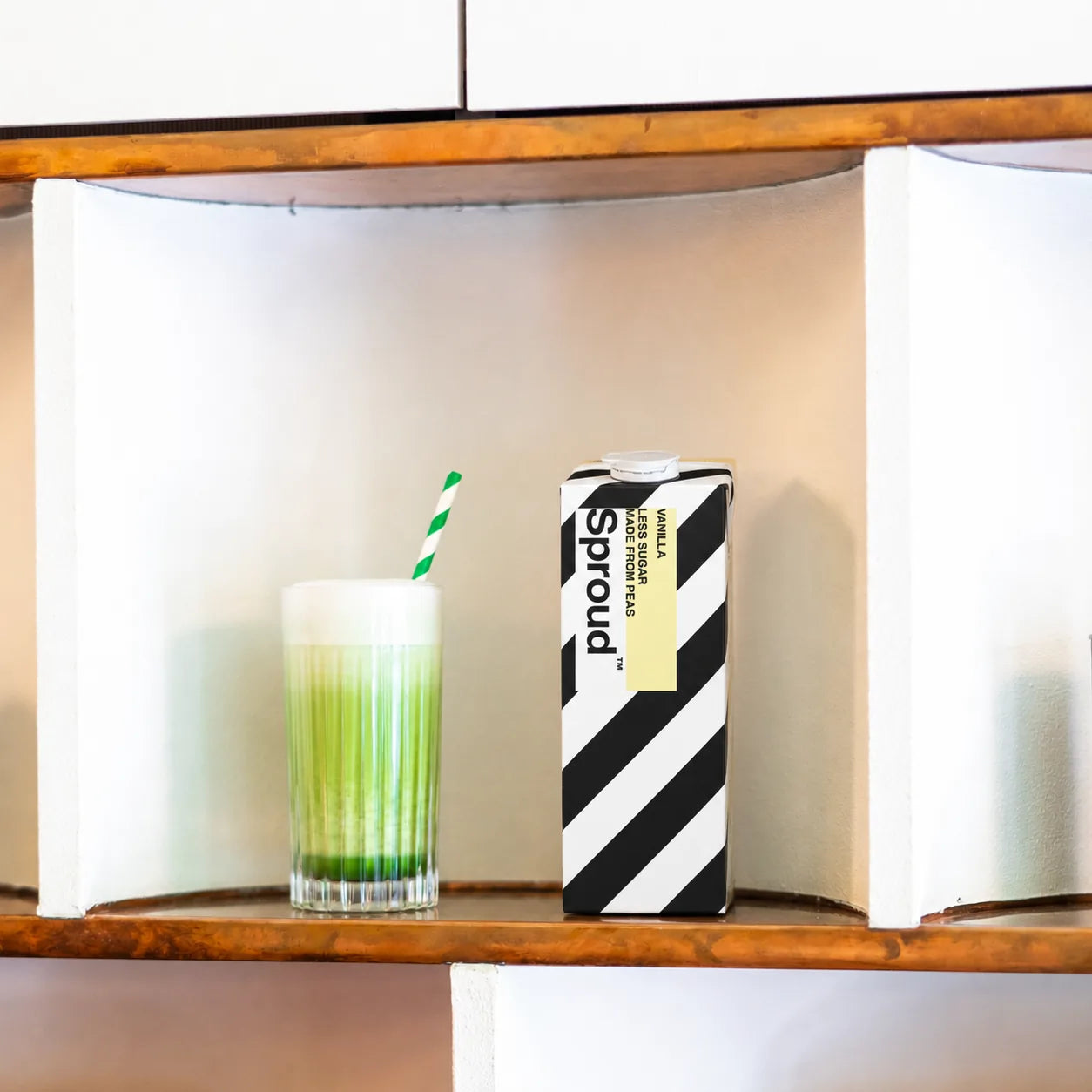 Matcha latte z mlekiem roślinnym Sproud Vanilla Less Sugar w szklance na drewnianej półce, zielona matcha z pianką i opakowanie Sproud Vanilla obok.