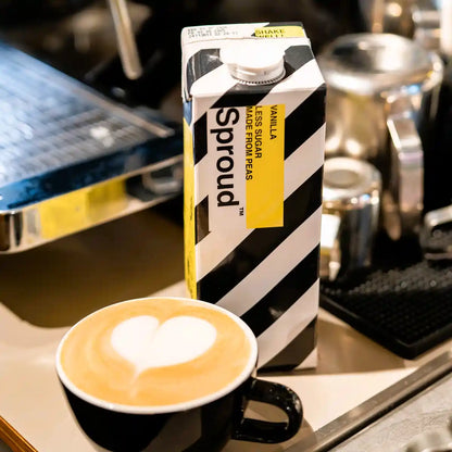 Karton napoju roślinnego Sproud Vanilla 1L obok filiżanki kawy z latte art – wegański napój z grochu o smaku waniliowym, idealny do kaw mlecznych.