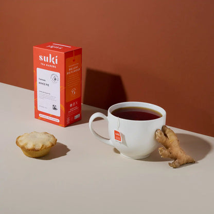 herbata Suki Tea Mince Pie Fairtrade w opakowaniu obok filiżanki herbaty, kawałka imbiru i ciastka mince pie na beżowo-brązowym tle