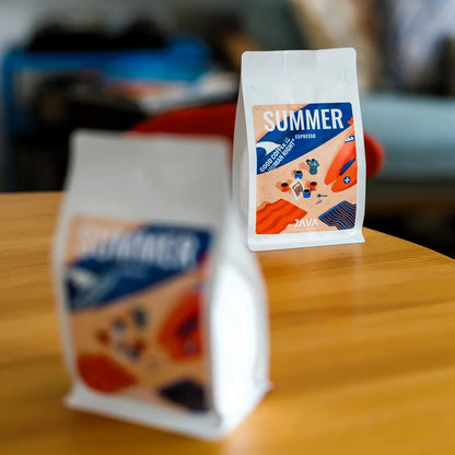 Dwie paczki Summer Espresso od JAVA Coffee Roasters ustawione na drewnianym stole z efektem bokeh i selektywną głębią ostrości
