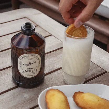 Osoba wkładająca ciastko do napoju limonkowego w wysokiej szklance, na drewnianym stole, obok butelki z aromatem waniliowym i talerza z ciastkami